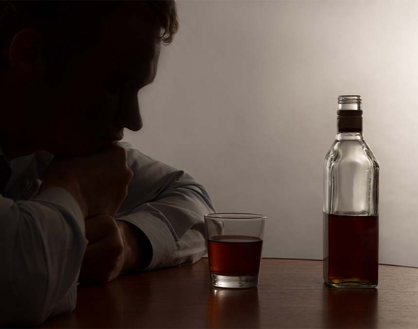 Hombre recargado en una mesa café, con un vaso y una botella de whisky