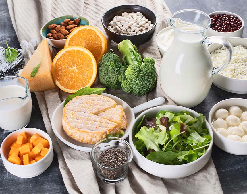 Diversos alimentos como leche, naranja, quesos, chía, brócoli, mango, melón, almendras y lechuga sobre una mesa negra