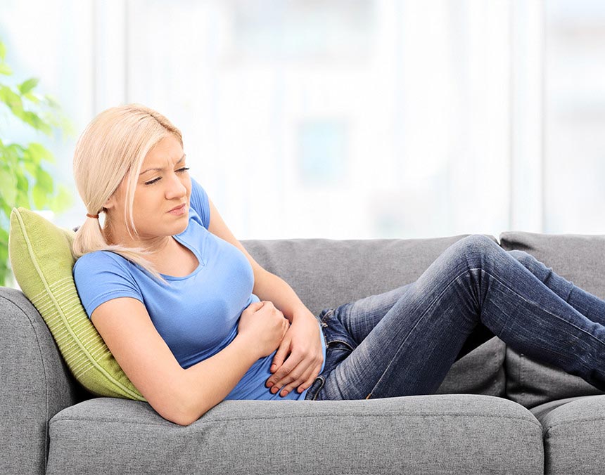 Mujer joven recostada en un sofá gris, tocando su abdomen y haciendo una expresión de dolor