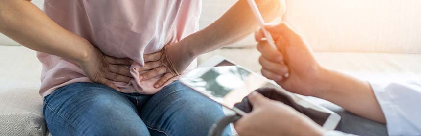 Mujer sentada en la camilla de un consultorio con dolor de vesícula, mientras es evaluada por un doctor quien sostiene un iPad
