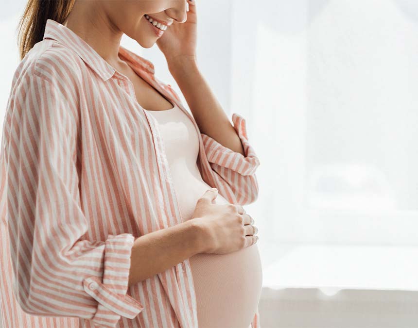 Mujer sonriendo, con siete meses de embarazo. Está vestida con una blusa blanca y camisa rosa