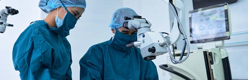 Dos médicos vestidos con pijamas azules para cirugía, gorros y cubrebocas, realizando una cirugía ocular