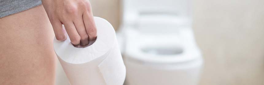 Mano de una mujer agarrando un rollo de papel higiénico. Al fondo está un retrete de color blanco