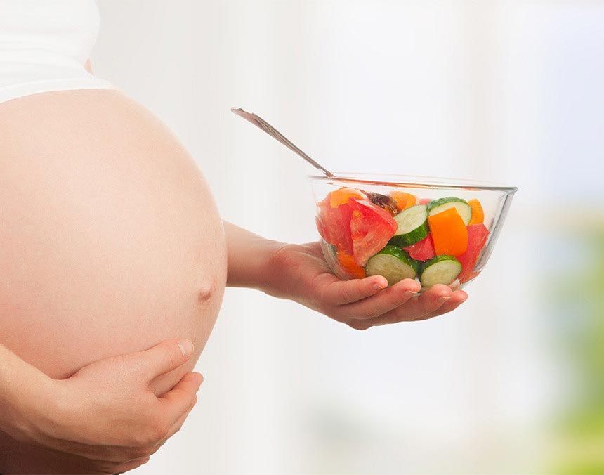 Mujer embarazada agarrándose el vientre con su mano derecha, mientras que con la izquierda está sosteniendo un tazón con verdura