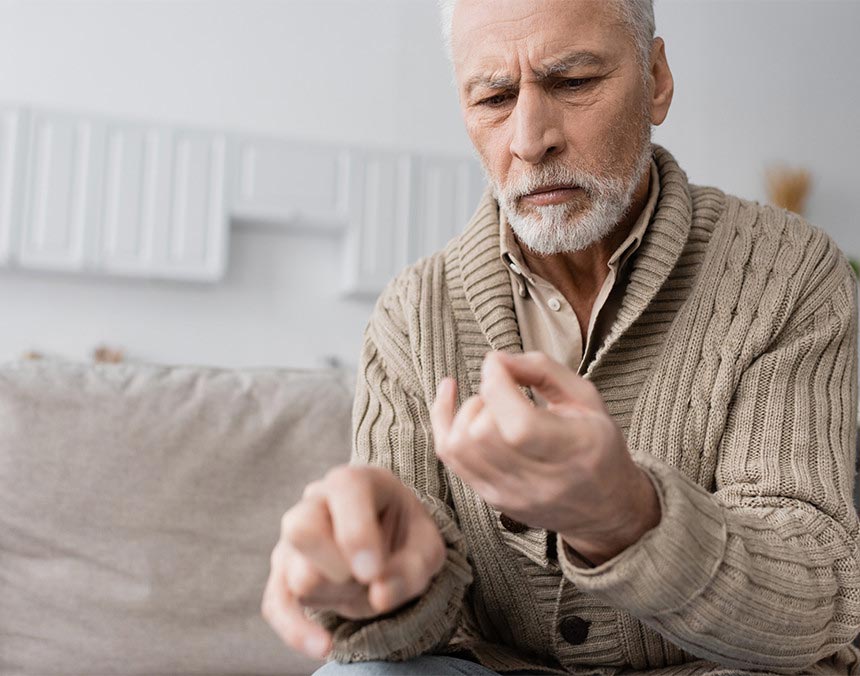 Hombre de la tercera edad sentado y mirando su mano que presenta rigidez a causa de la enfermedad de Parkinson