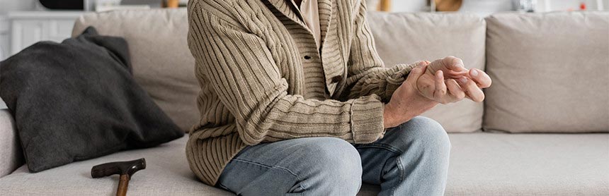 Hombre de la tercera edad sentado en un sofá, agarrando su mano que presenta rigidez a causa de la enfermedad de Parkinson