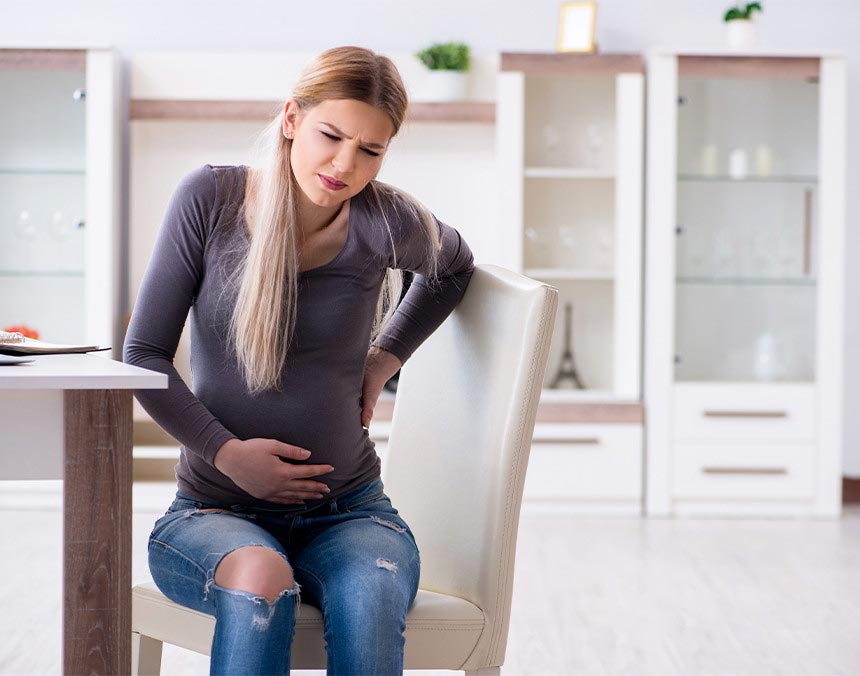 Mujer embarazada sentada en una silla del comedor. Está agarrándose el abdomen y la espalda, mostrando malestar