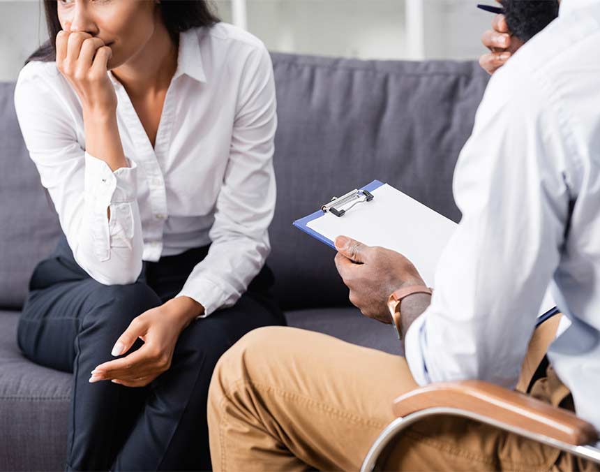 Paciente vestida con camisa blanca y pantalón negro, sentada en el sofá de un consultorio mientras está en cita con un psicólogo y él toma notas