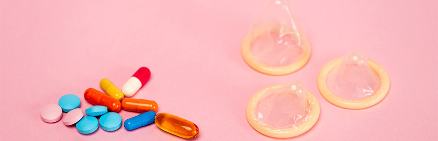 Pastillas anticonceptivas de color azul, naranja, amarillo, rojo y blanco, y tres condones masculinos, sobre una superficie rosa
