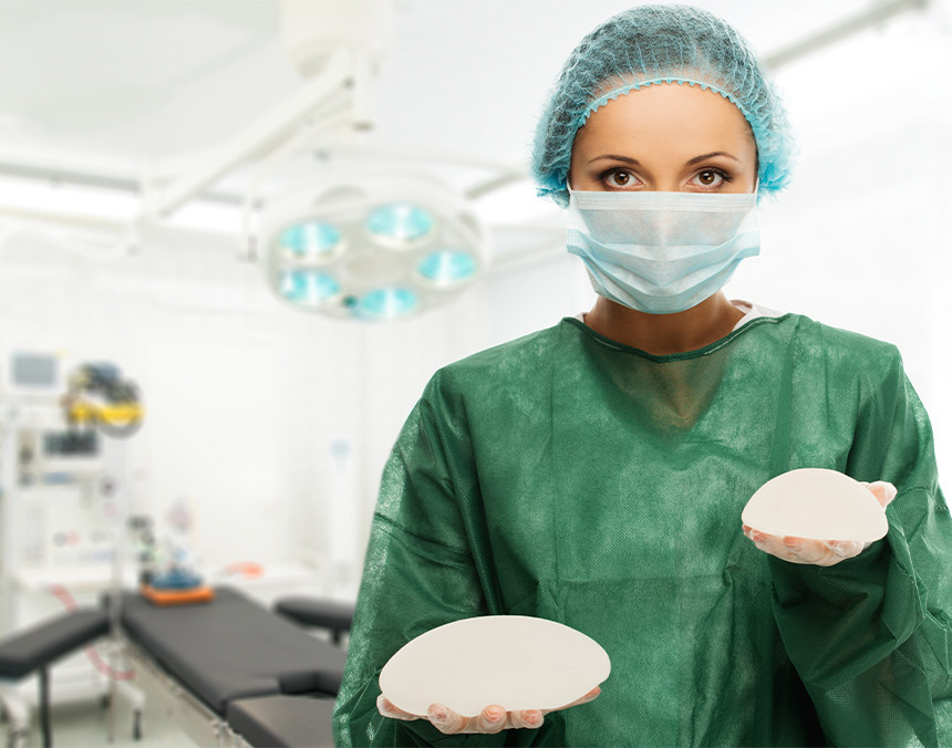 Doctora vestida con pijama para cirugía, guantes blancos y gorro, sosteniendo dos implantes mamarios. Al fondo hay una camilla quirúrgica