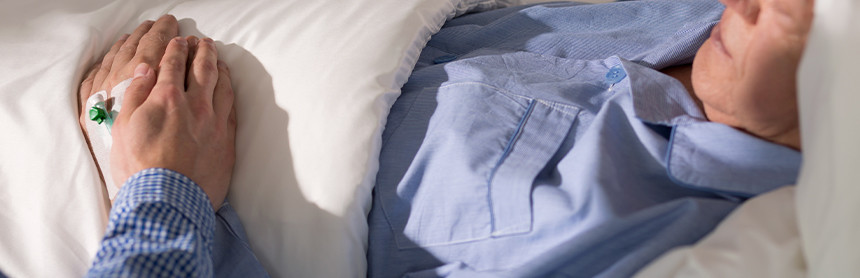 Hombre de la tercera edad acostado en una cama dormido y con el suero intravenoso, mientras un familiar le sostiene la mano