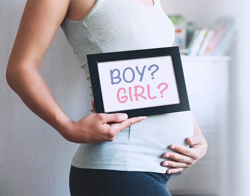 Mujer embarazada vestida con playera gris y licra negra, sosteniendo un cartel que dice ¿niño o niña?