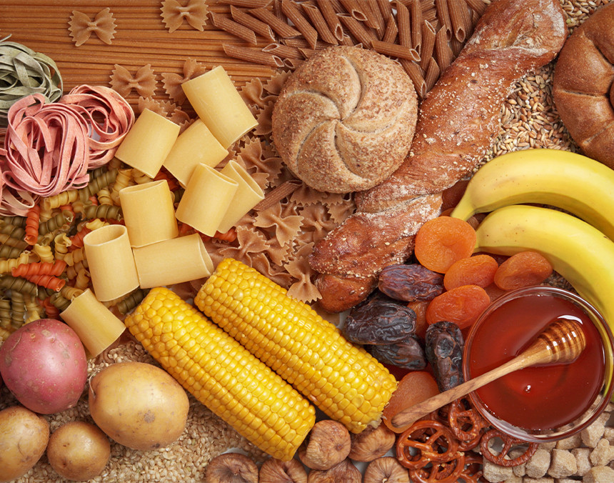 Diversos tipos de pasta cruda, pan, cereales, frutos secos, plátanos, elotes amarillos y miel sobre una superficie de madera