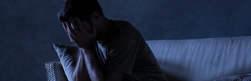 Es de noche. Un hombre está sentado en un sofá con las manos en su cara, mostrando desesperación