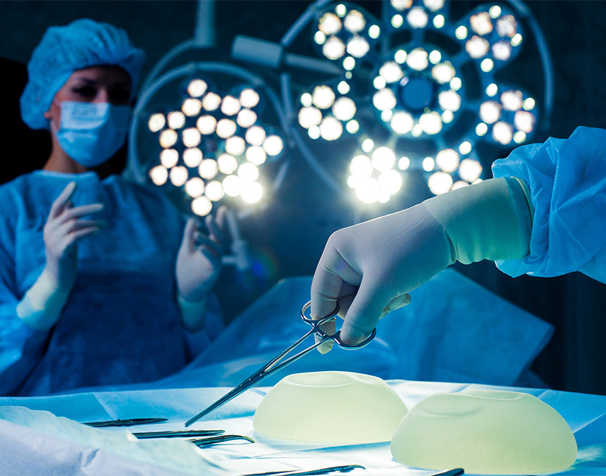 Dos médicos vestidos con pijamas azules, gorro y cubrebocas. Uno sostiene una pinza y a su lado hay dos implantes mamarios