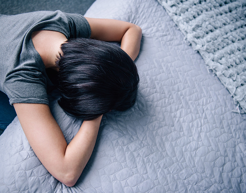 Mujer joven hincada de rodillas y recargada en una cama cubierta con una colcha gris. Sus manos están bajo su cabeza
