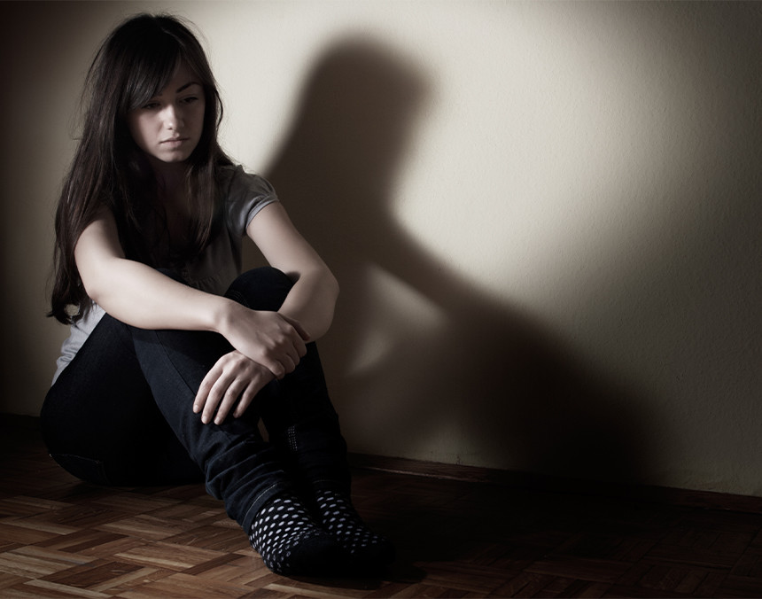 Adolescente deprimida sentada en el suelo. Esta vestida con una playera gris y pantalón negro