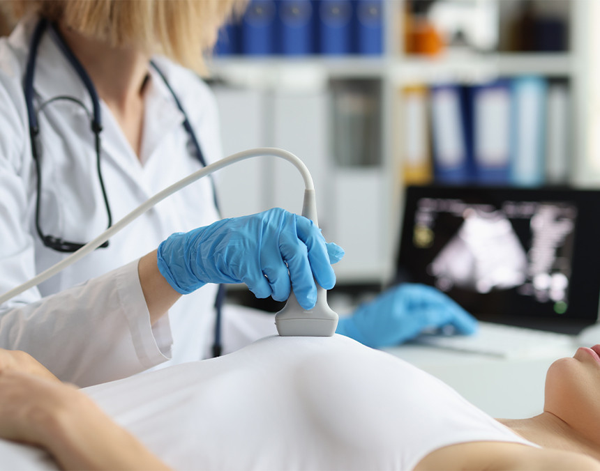 Doctora con bata blanca y guantes azules realizando un ultrasonido mamario a una paciente que está recostada