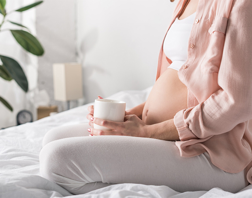Mujer joven embarazada sentada en una cama y sosteniendo una taza blanca. Viste con una blusa rosa y pants grises