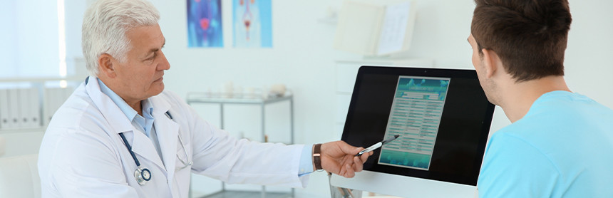 Doctor con bata blanca señalando una computadora con una pluma mientras un paciente lo escucha con atención