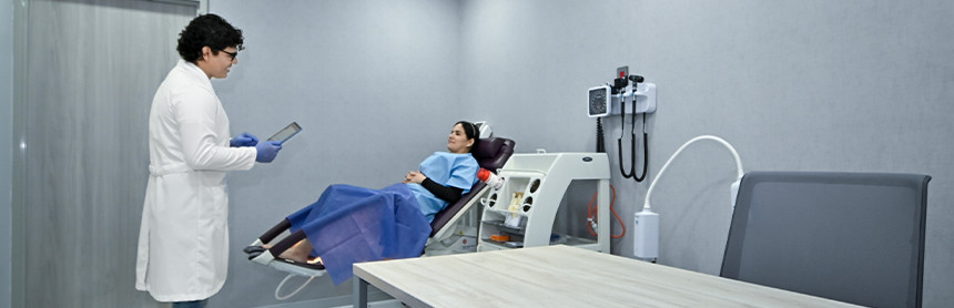 Médico con bata blanca y guantes azules frente a una paciente que está recostada en una silla de examen ginecológico