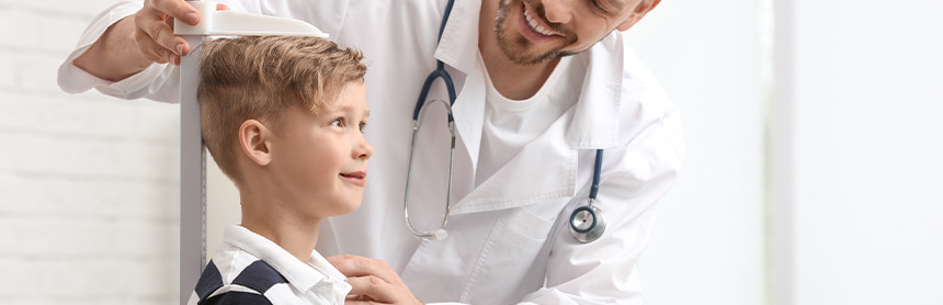 Médico con bata blanca midiendo la altura de un niño pequeño vestido con una playera de rayas negras y blancas