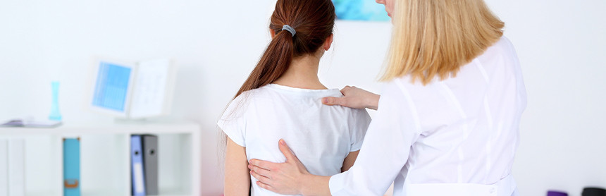 Fisioterapeuta con bata blanca examinando la espalda de una paciente que está sentada en la mesa de exploración