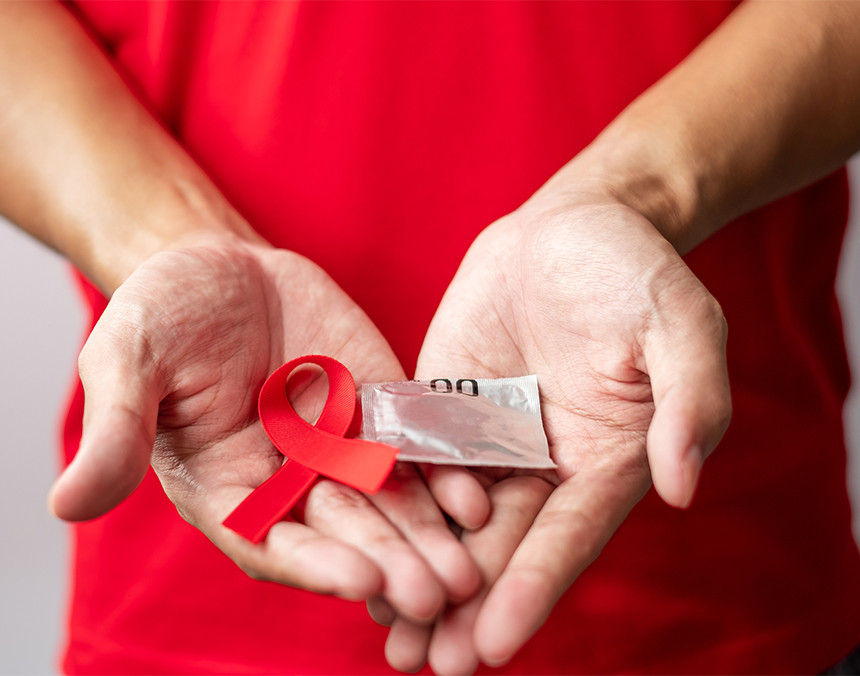 Hombre vestido con playera roja sosteniendo una cinta roja haciendo referencia a la lucha contra el SIDA y un preservativo