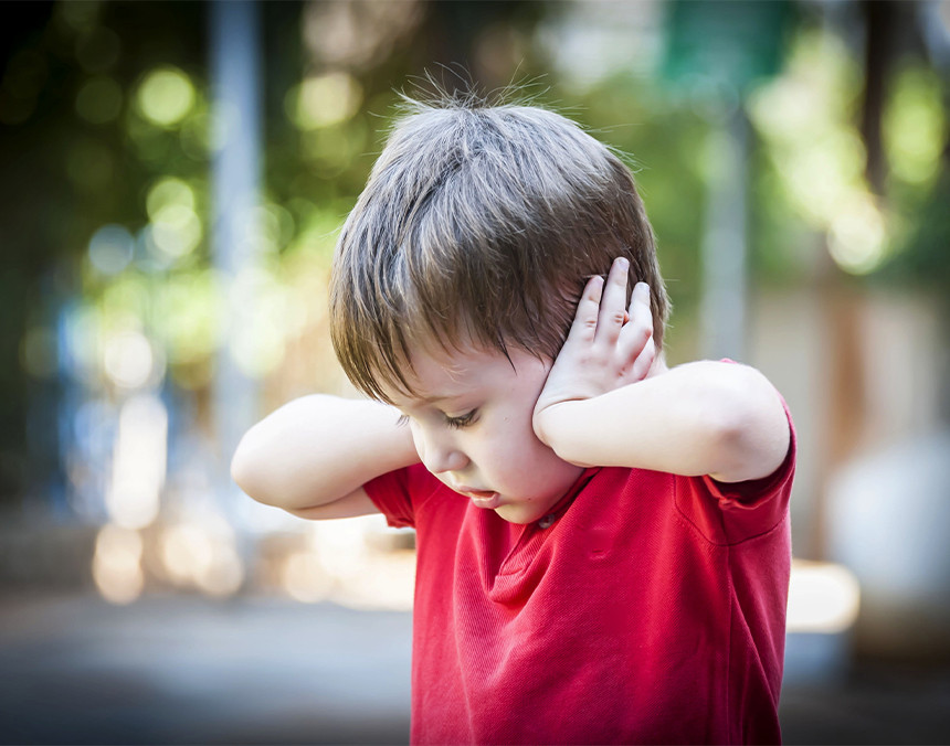 Niño pequeño vestido con playera roja tapándose los oídos con las manos. Está en un parque