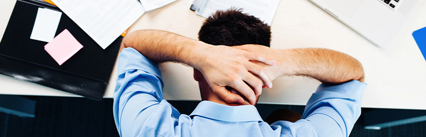 Hombre joven con estrés acostado en su escritorio. A lado de él hay varios papeles de oficina y una computadora