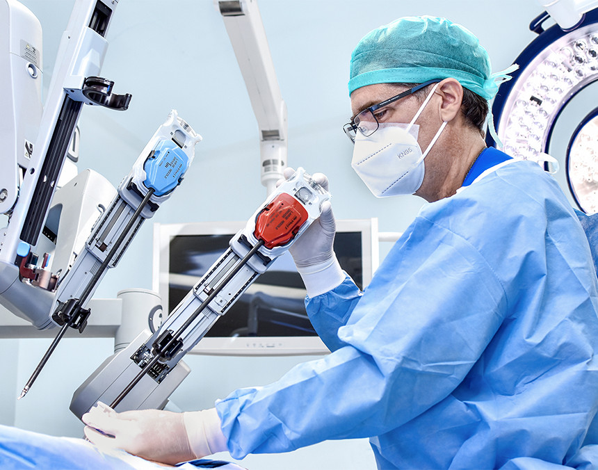 Médico con bata azul, gorro verde y cubrebocas realizando un procedimiento quirúrgico con el Sistema Quirúrgico Robótico Da Vinci