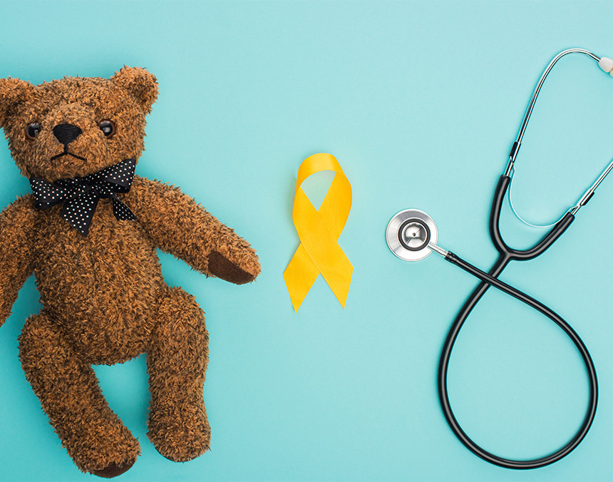 Peluche de un oso café, a lado un moño amarillo haciendo alusión al cáncer infantil y un estetoscopio sobre un fondo azul
