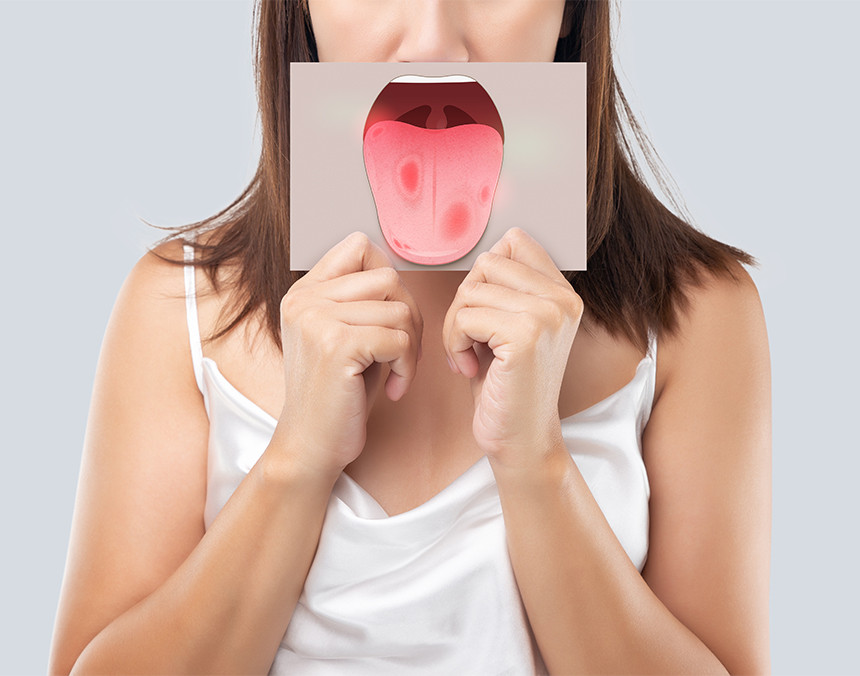 Mujer joven vestida con blusa blanca sosteniendo un cartel pequeño a la altura de su boca que muestra una lengua con cáncer