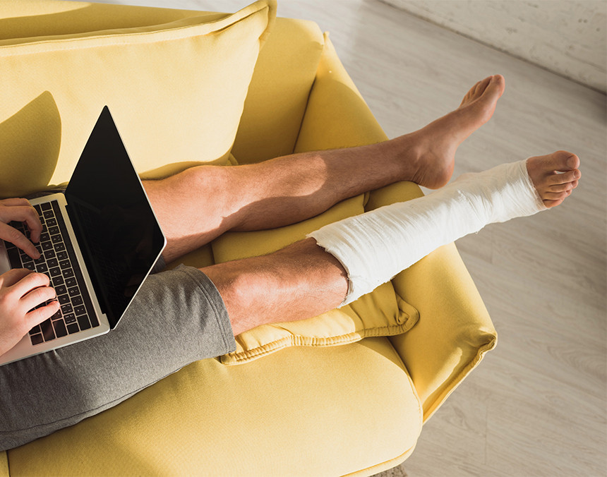 Hombre con una pierna enyesada recostado en un sofá de color amarillo y sosteniendo una computadora