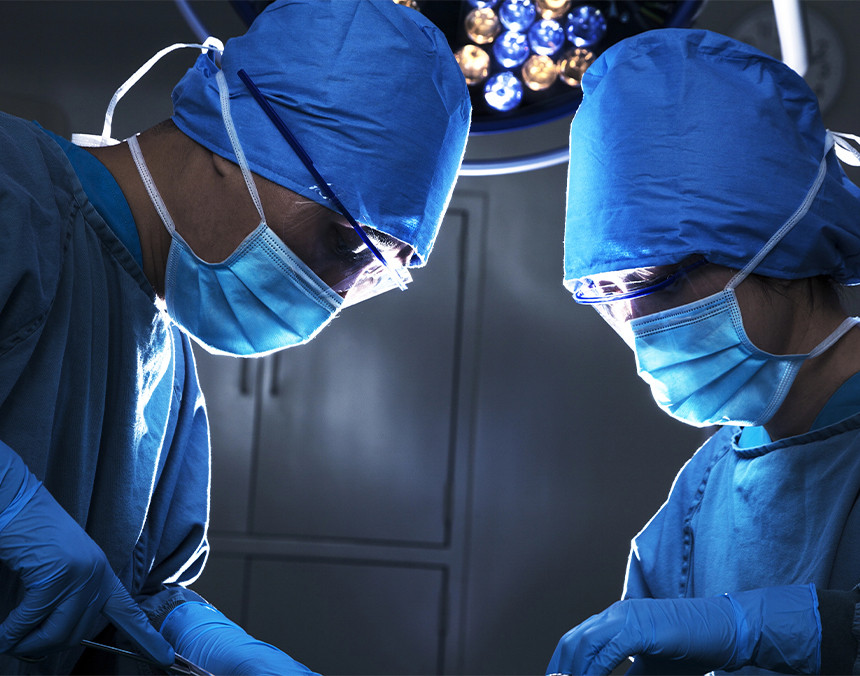 Dos médicos con pijamas azules para cirugía, gorros y cubrebocas, concentrados realizando un procedimiento quirúrgico