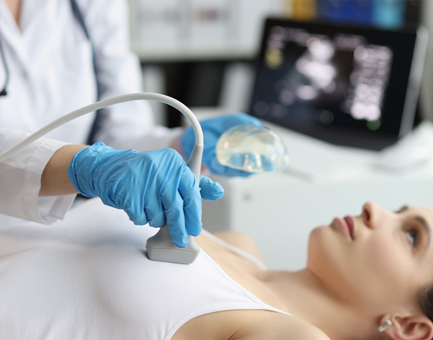 Mujer joven que viste una blusa sin mangas de color blanco está acostada en una camilla de observación mientras una doctora le realiza un ultrasonido mamario
