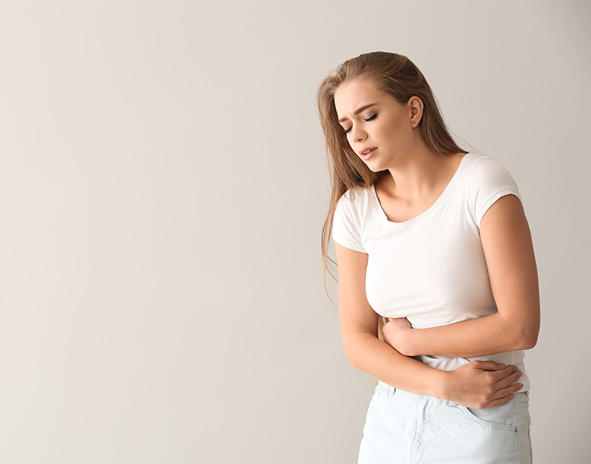 Mujer joven vestida con una blusa blanca y pantalón gris colocando sus manos en su vientre a causa del dolor