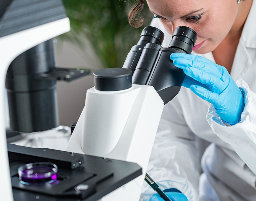 Doctora con bata blanca y guantes azules analizando una muestra de laboratorio con un microscopio