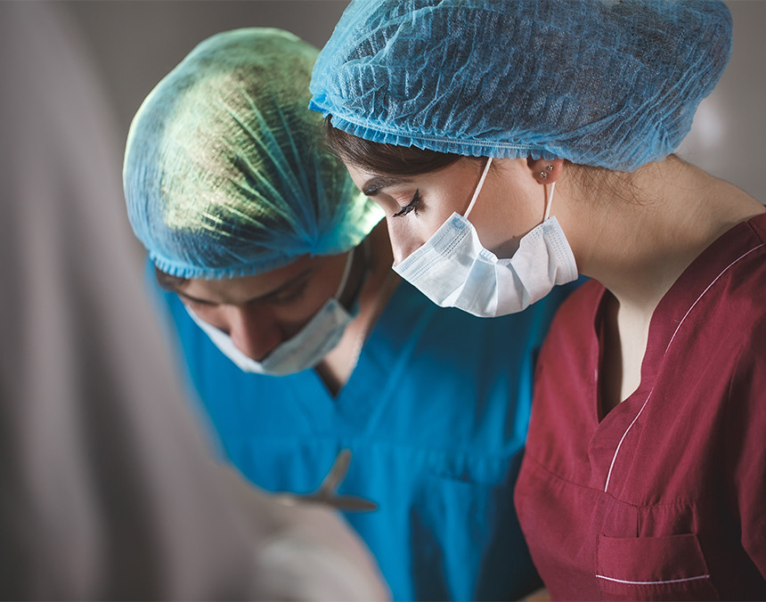 Dos doctoras vestidas con pijamas para cirugía, gorros y cubrebocas, realizando un procedimiento quirúrgico