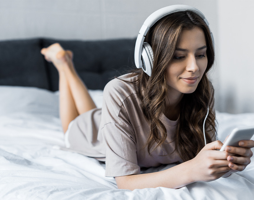 Mujer joven acostada en una cama, escuchando música con sus audífonos y viendo su teléfono