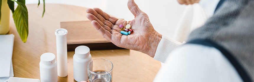 Persona de la tercera edad sosteniendo unas pastillas en su mano. A lado hay un vaso de agua