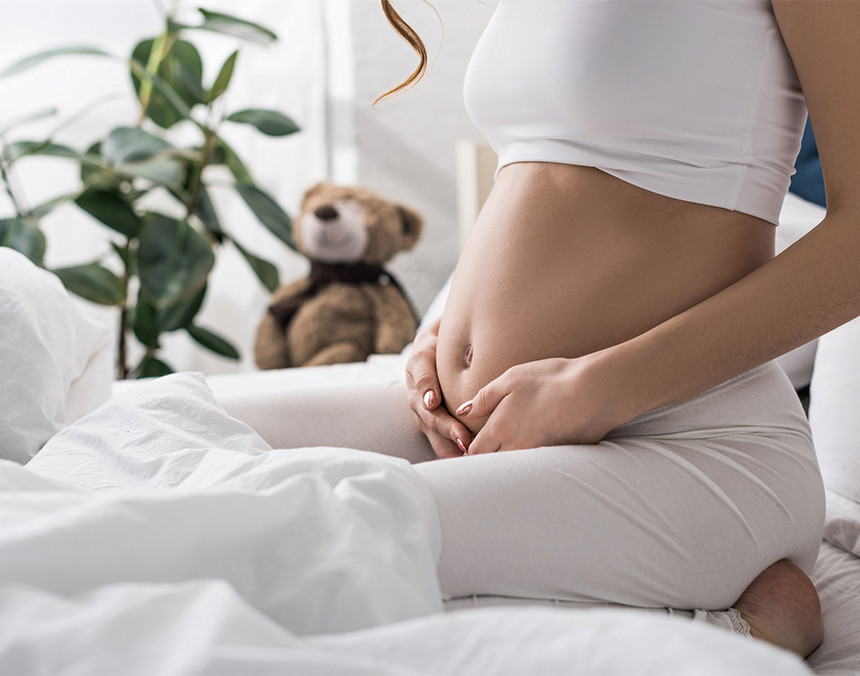 Mujer embarazada de rodillas sobre una cama con sábanas blancas. Al fondo hay un oso de peluche