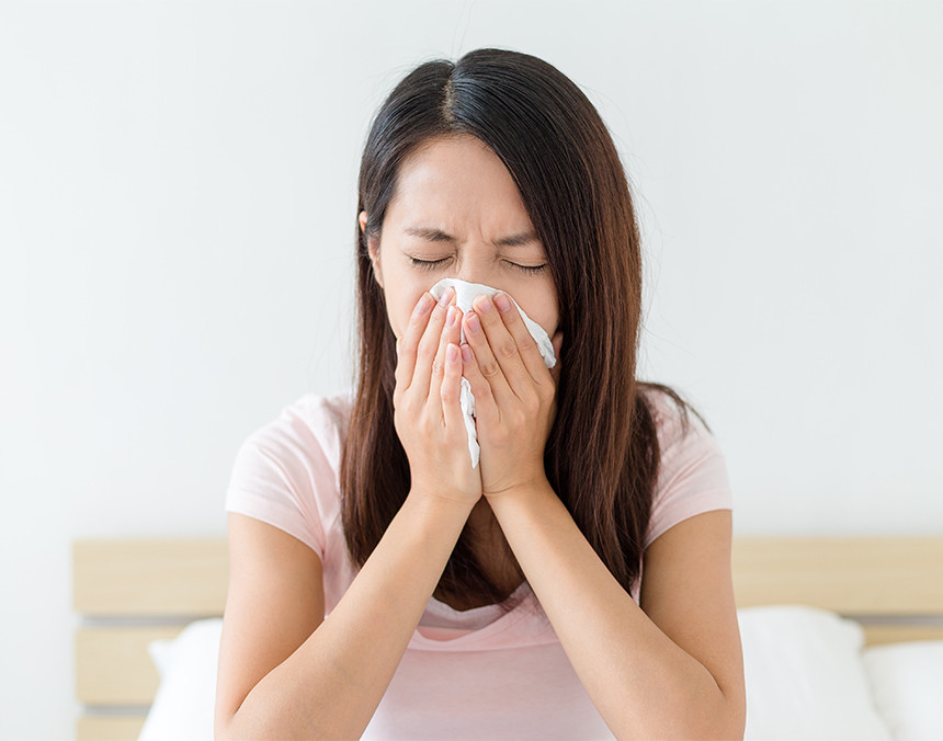Mujer joven con alergias está sentada en una cama sonándose la nariz con un pañuelo desechable