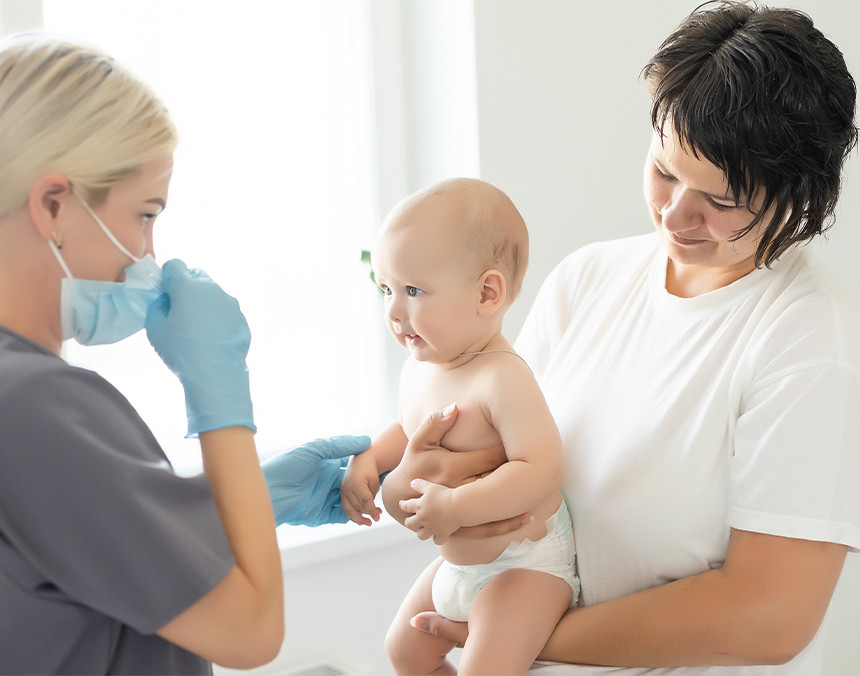 Doctora vestida con uniforme gris y cubrebocas, vacunando a un bebé de pocos meses de edad en un consultorio
