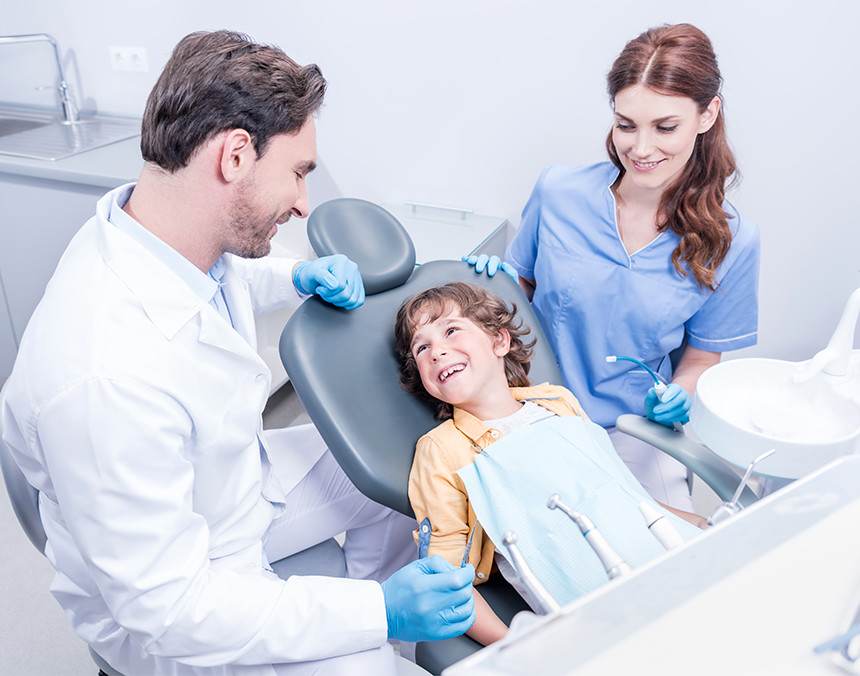 Niño sentado en una silla de dentista, sonriendo hacia un médico odontólogo que usa bata blanca y guantes azules. La madre se encuentra a lado del niño