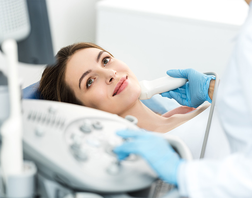 Médico con bata blanca y guantes azules examinando la glándula tiroidea de una paciente que está recostada en una mesa de observación
