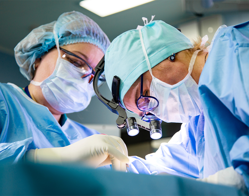 Dos médicos vestidos con pijamas azules para cirugía, guantes y cubrebocas, realizando un procedimiento quirúrgico