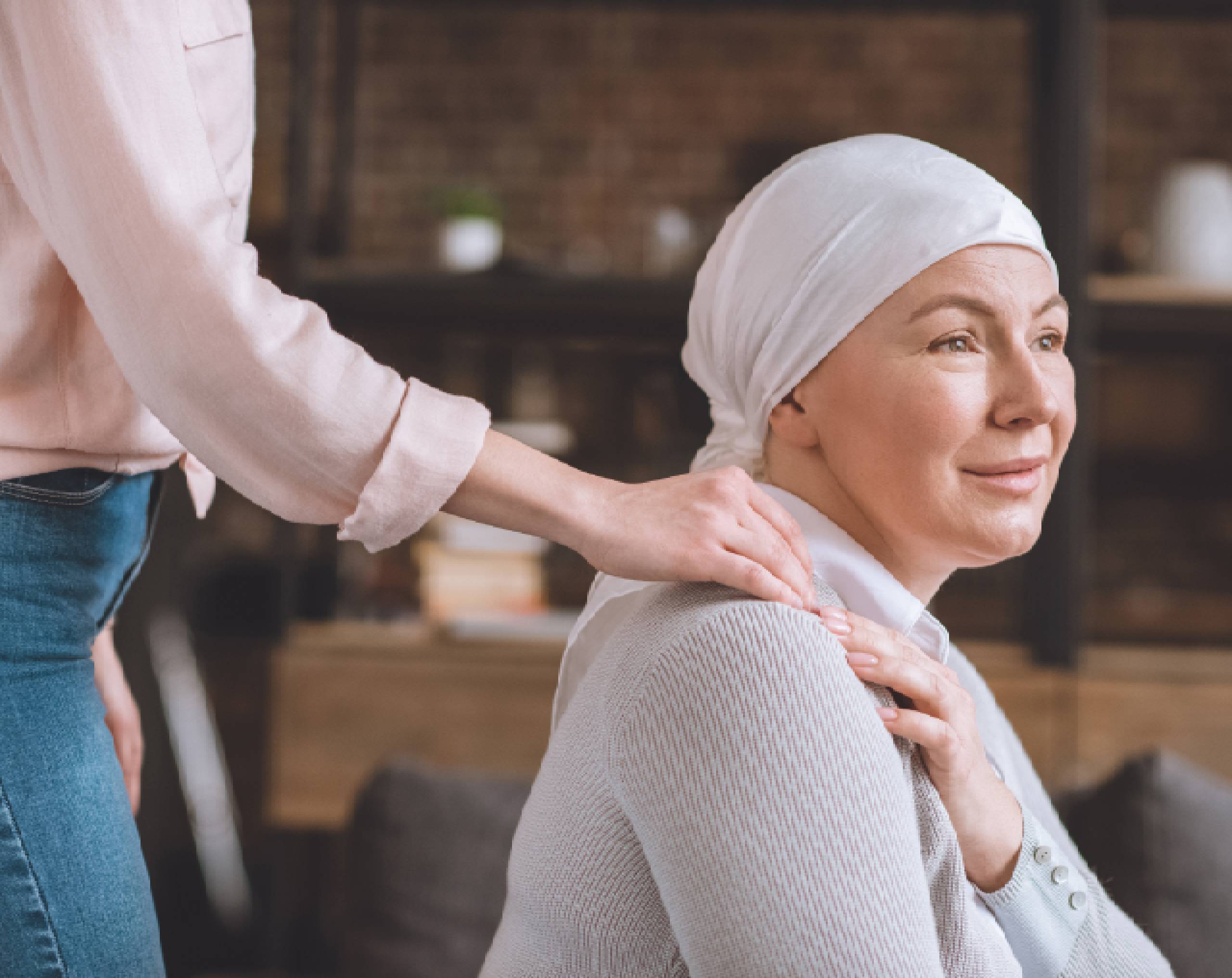 Mujer con cáncer sentada usa un pañuelo en la cabeza y una mujer le toca el hombro por otra mujer en señal de apoyo.