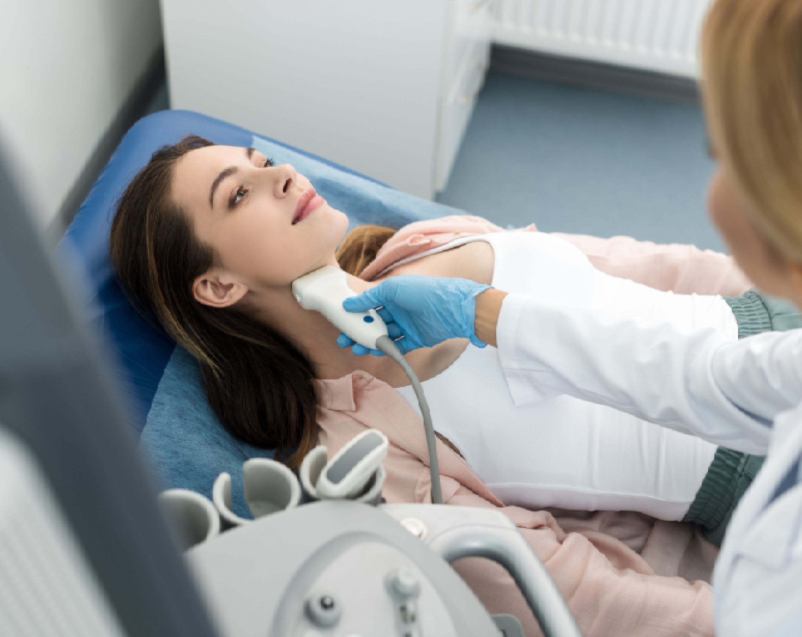 Mujer de edad media está recostada en el consultorio y una doctora le hace un ultrasonido de tiorides para detectar hipotiroidismo.