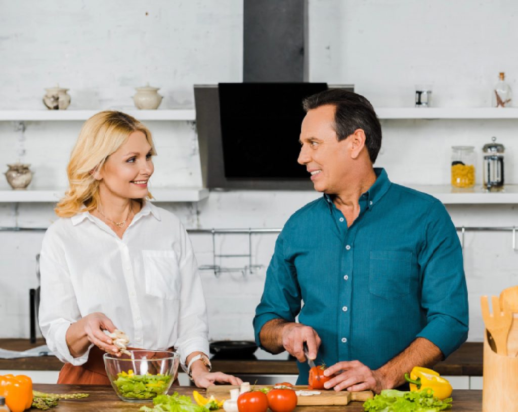 Pareja de edad media se miran con complicidad mientras cocinan comida saludable en la cocina, previniendo la prediabetes.
